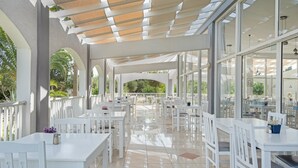 Un espacio comedor exterior techado con mesas y sillas blancas, ventanas amplias y vistas a la vegetación.