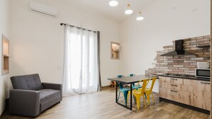 Un salon moderne comprenant un canapé, une table à manger, des chaises et un coin cuisine avec un mur de fond en briques et des appareils électroménagers.