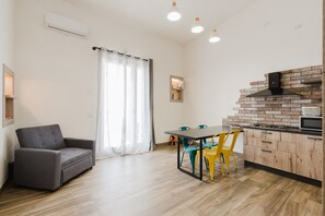 A modern living room with a sofa, dining table, chairs, and a kitchen area with a brick backsplash and appliances.