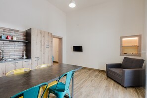 A modern kitchen with a wooden backsplash, a dining table with chairs, a grey sofa, and a flat-screen TV mounted on the wall.