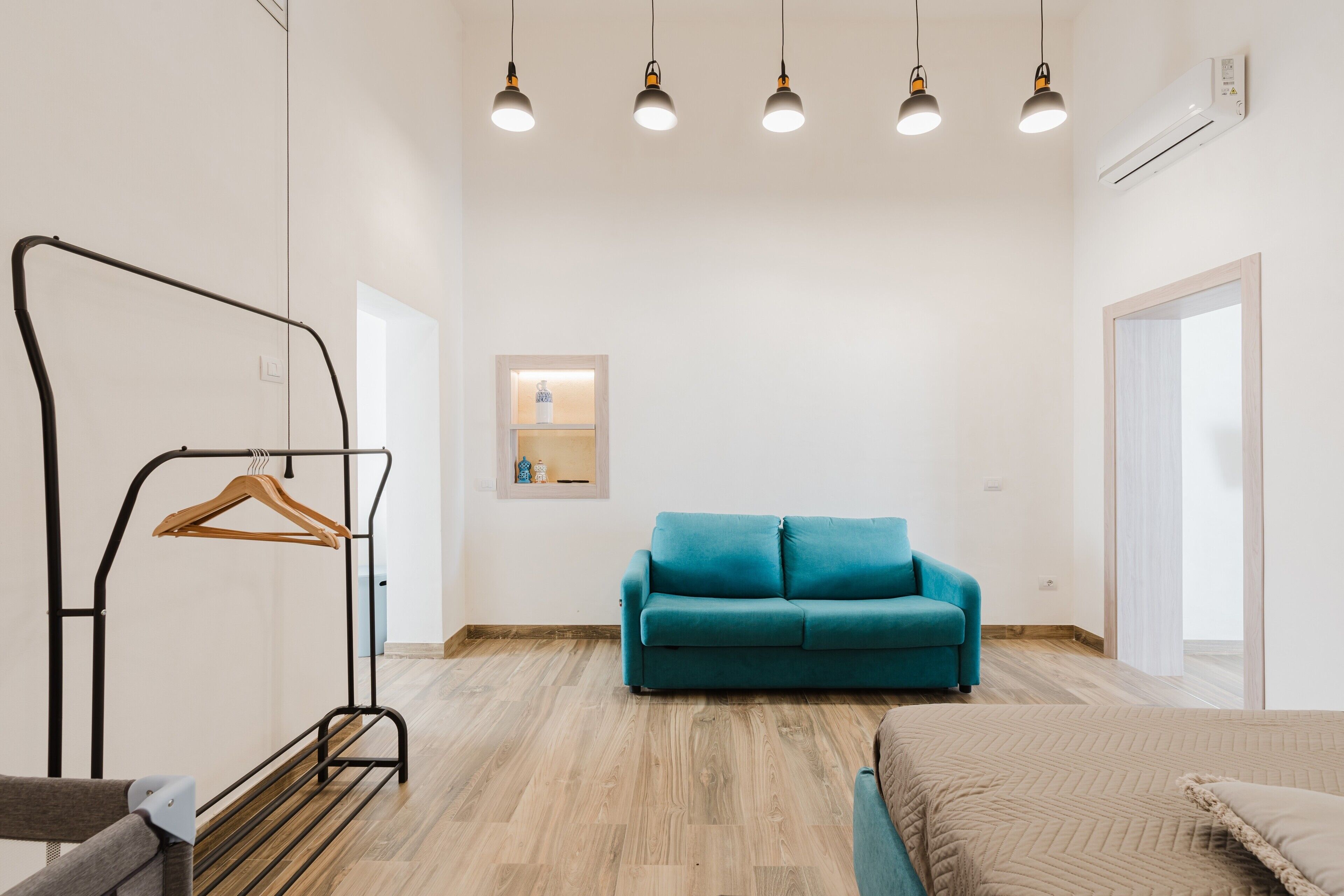 Una habitación con una cama, un sofá azul, suelo de madera, una lámpara colgante y un aire acondicionado instalado en la pared.