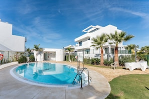 Un'area piscina moderna con vasca azzurra e cristallina, circondata da palme e un edificio bianco con balconi.