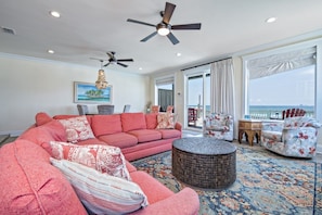 A spacious living room with a large sofa, a round ottoman, and two armchairs. There are ceiling fans, a chandelier, and a large window with curtains.