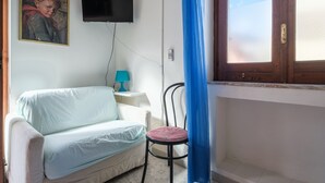 Ein Zimmer mit einem weißen Sofa, einem kleinen Tisch mit Lampe, einem an der Wand befestigten Fernseher und einem Fenster mit blauer Vorhang.