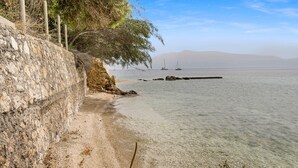 Paesaggio costiero con un muro di pietra, acqua limpida e velieri in lontananza.