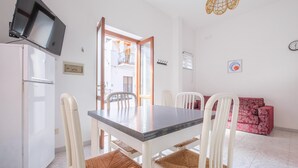 Un área de comedor con una mesa y sillas, un televisor montado en la pared, un sofá rojo con estampado, y un balcón visible a través de una puerta abierta.