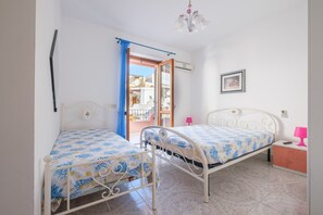 Un dormitorio con dos camas de metal, una ventana con cortinas azules, una lámpara rosa y un cuadro en la pared.