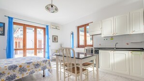 A room with a bed, a dining table with chairs, a kitchen area with white cabinets, and a window with blue curtains.