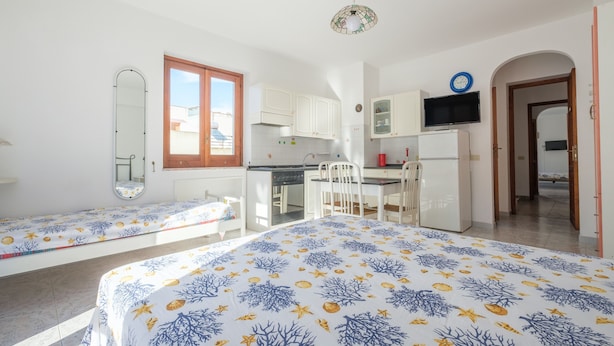 A room with a bed, a desk, a chair, a mirror, a television, and a kitchen area.