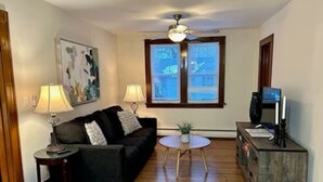 A living room with a black sofa, two side tables with lamps, a small round table, a television on a wooden stand, and a window with blinds.