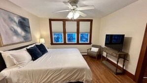 A bedroom with a bed, a ceiling fan, a TV, a chair, and a window with blinds.