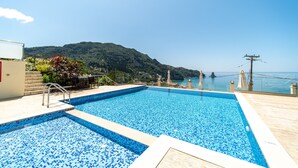 A rectangular swimming pool with blue mosaic tiles, a wooden deck, and a view of a mountainous landscape. - Studio Apartment "Mazis Studio B2" with Sea View, Balcony & Wi-Fi (Corfu)