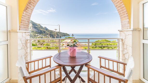 Un balcon avec une table et des chaises en bois, un vase rose, et une vue sur la mer et la verdure.
