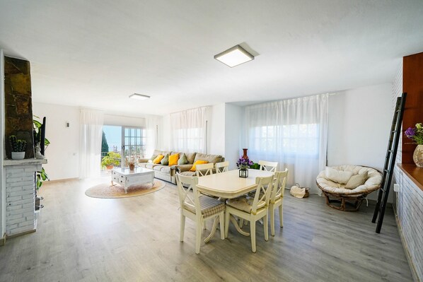 A spacious living area with a dining table, chairs, a sofa, and a large window.