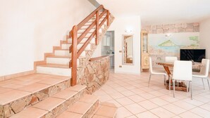 A staircase with wooden railings leading to a stone wall, a dining area with a table and chairs, and a mural on the wall. - Holiday Home 'Villa Alfredo Golfo Aranci' with Sea View, Private Terrace, and Air Conditioning (GolfoAranci)