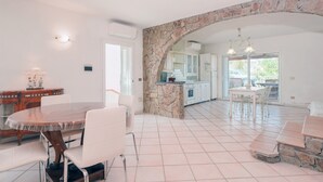 A spacious kitchen with a stone archway, white cabinetry, and a dining area with a round table and chairs. - Holiday Home 'Villa Alfredo Golfo Aranci' with Sea View, Private Terrace, and Air Conditioning (GolfoAranci)
