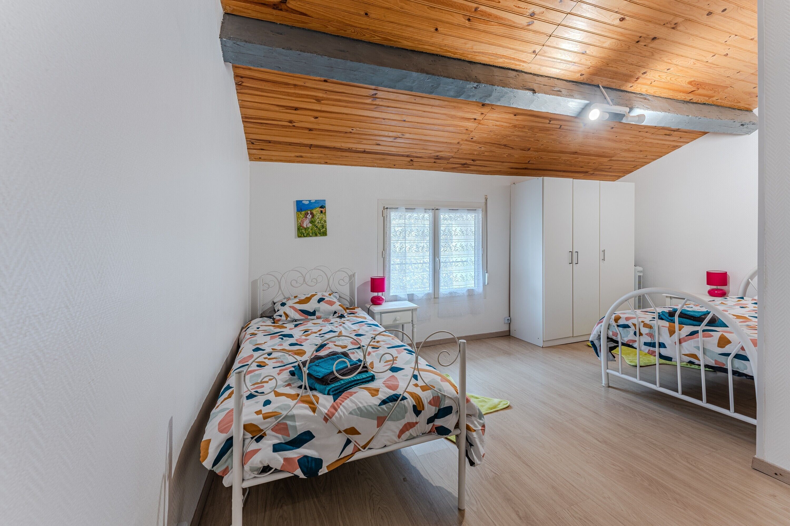 Une chambre avec deux lits, des poutres apparentes en bois au plafond, une fenêtre avec des stores et une petite table de chevet avec une lampe.