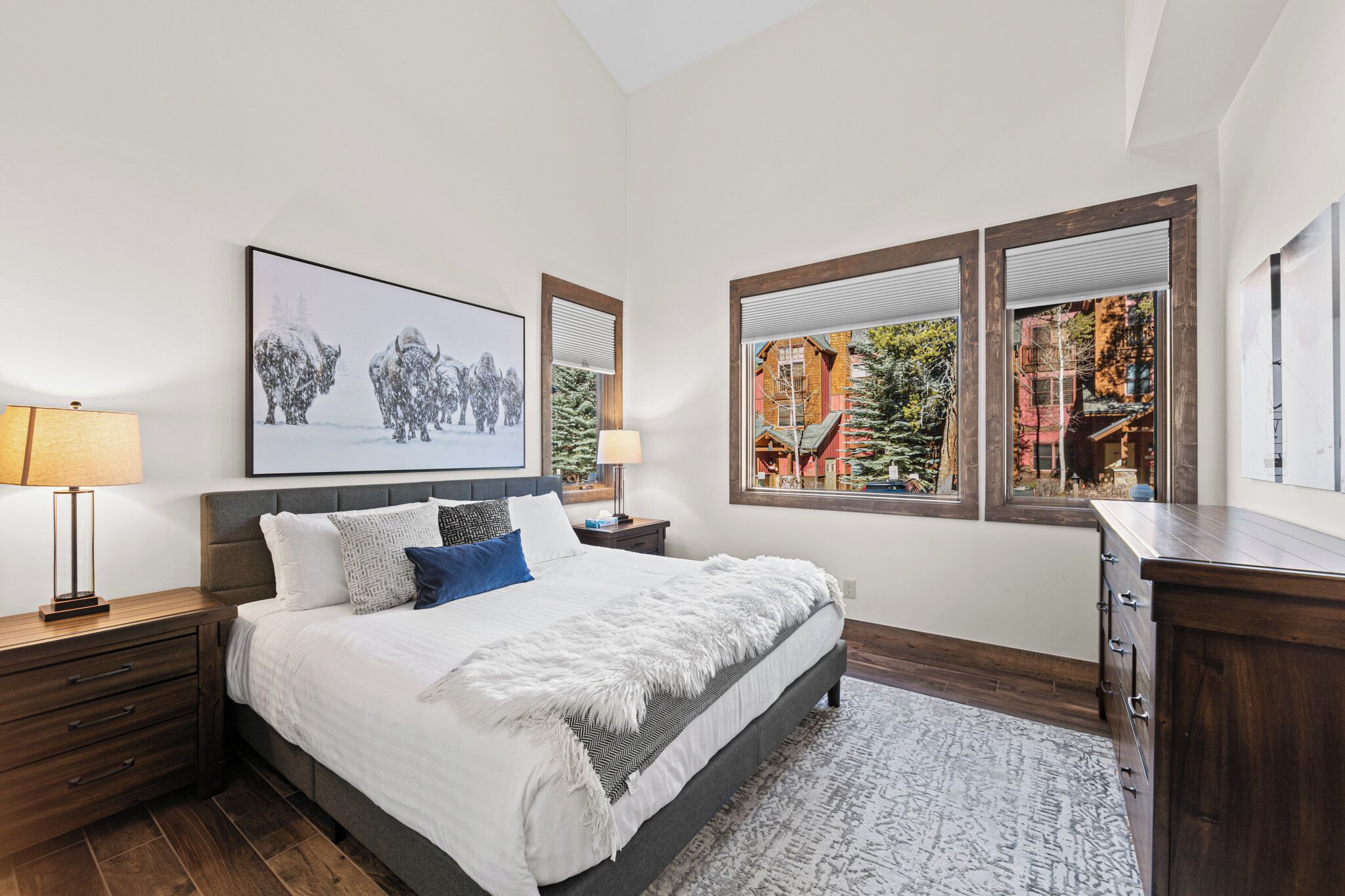 A bedroom with a large bed, two bedside lamps, a framed picture, and a window with a view of a building and trees.