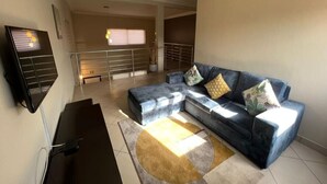 A living room with a sectional sofa, a flat-screen TV mounted on the wall, and a staircase leading to an upper floor. - 🌴 Your Dream Getaway in the heart of Accra! Stay in this 3 Bedroom Property! 🏡 (Accra)