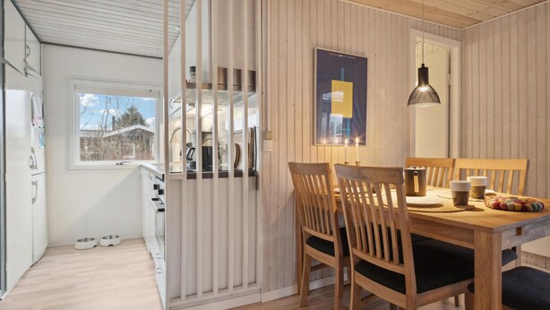 Une cuisine moderne avec des meubles blancs, un coin repas avec des chaises et une table en bois, et une vue sur l’extérieur par une fenêtre.