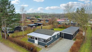 Un bâtiment moderne de style cabane, doté d’un grand garage et d’un porche abrité, entouré d’un paysage rural avec des maisons et des arbres.