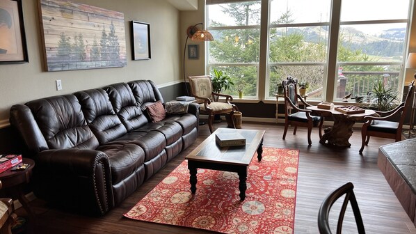 A living room with a leather sofa, a coffee table, and a view of greenery through large windows. - Secluded getaway 5 minutes from salmon fishing, kayaking, and whale watching! (Curry County)