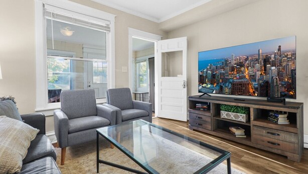 Un salon moderne avec une table basse en verre, des canapés gris, un tableau représentant un paysage urbain et un meuble de rangement en bois pour les appareils électroniques.