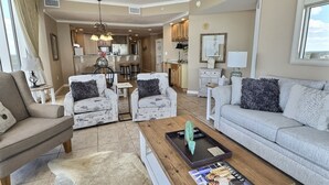 A spacious living room with a sofa, armchairs, and a coffee table. A kitchen area is visible in the background. - Top of The World * Penthouse Views * Free Daily Activities! (Gulf Shores)