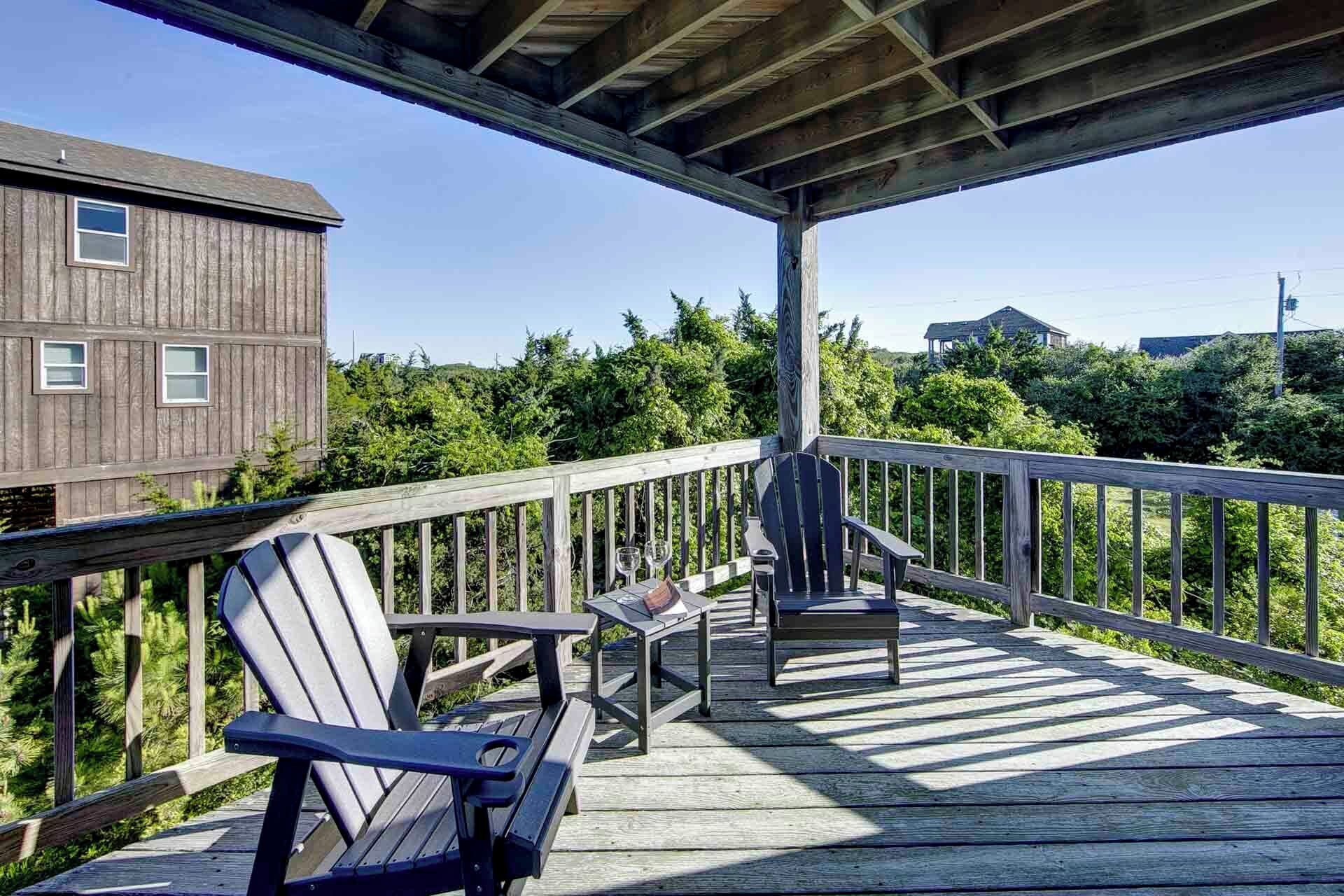 A wooden deck with Adirondack chairs and a table, overlooking a forested area.