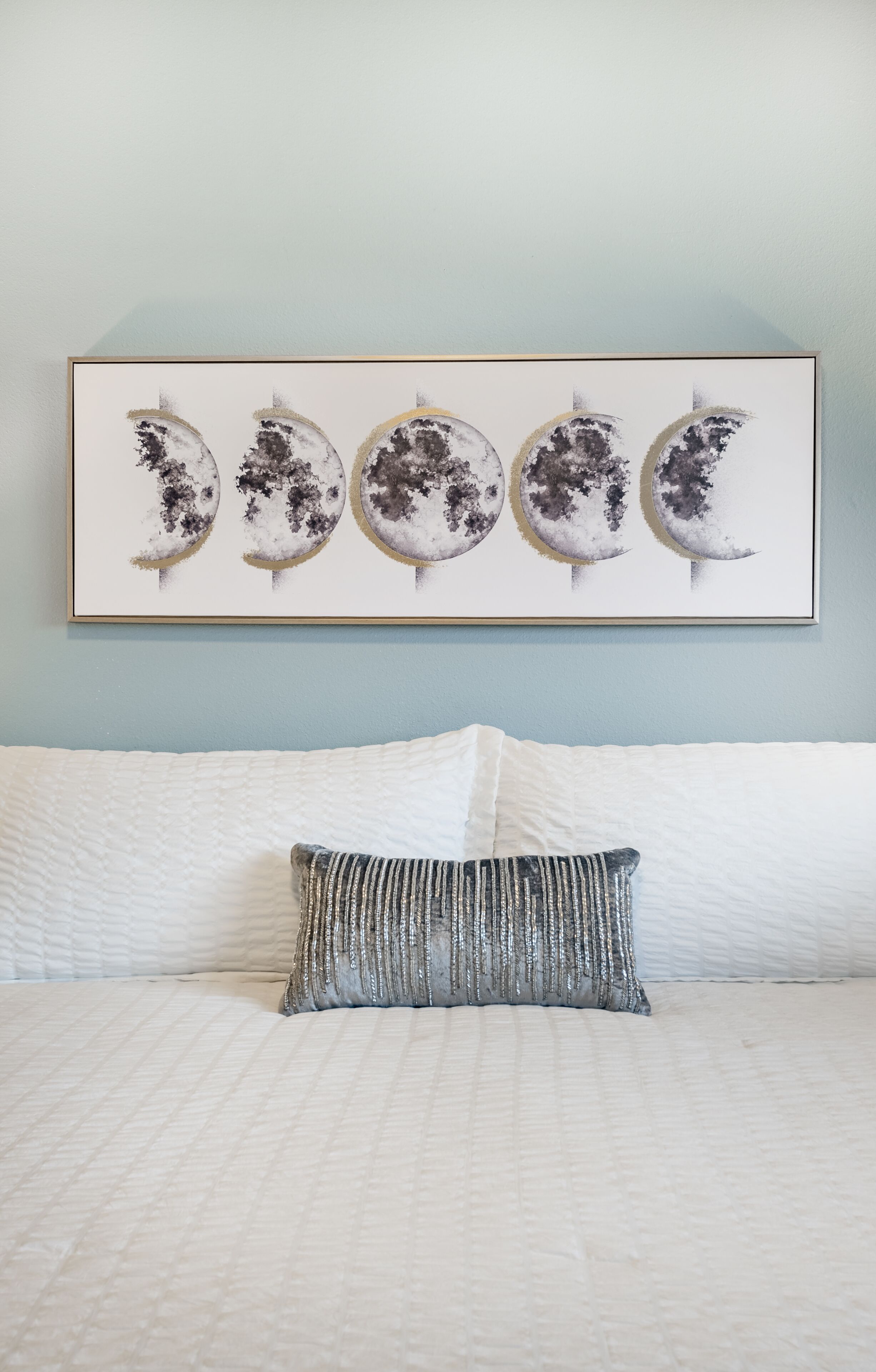 Une œuvre d’art encadrée représentant les quatre phases de la lune, accrochée au mur au-dessus d’un lit recouvert d’une literie blanche et agrémenté d’un coussin à texture particulière.