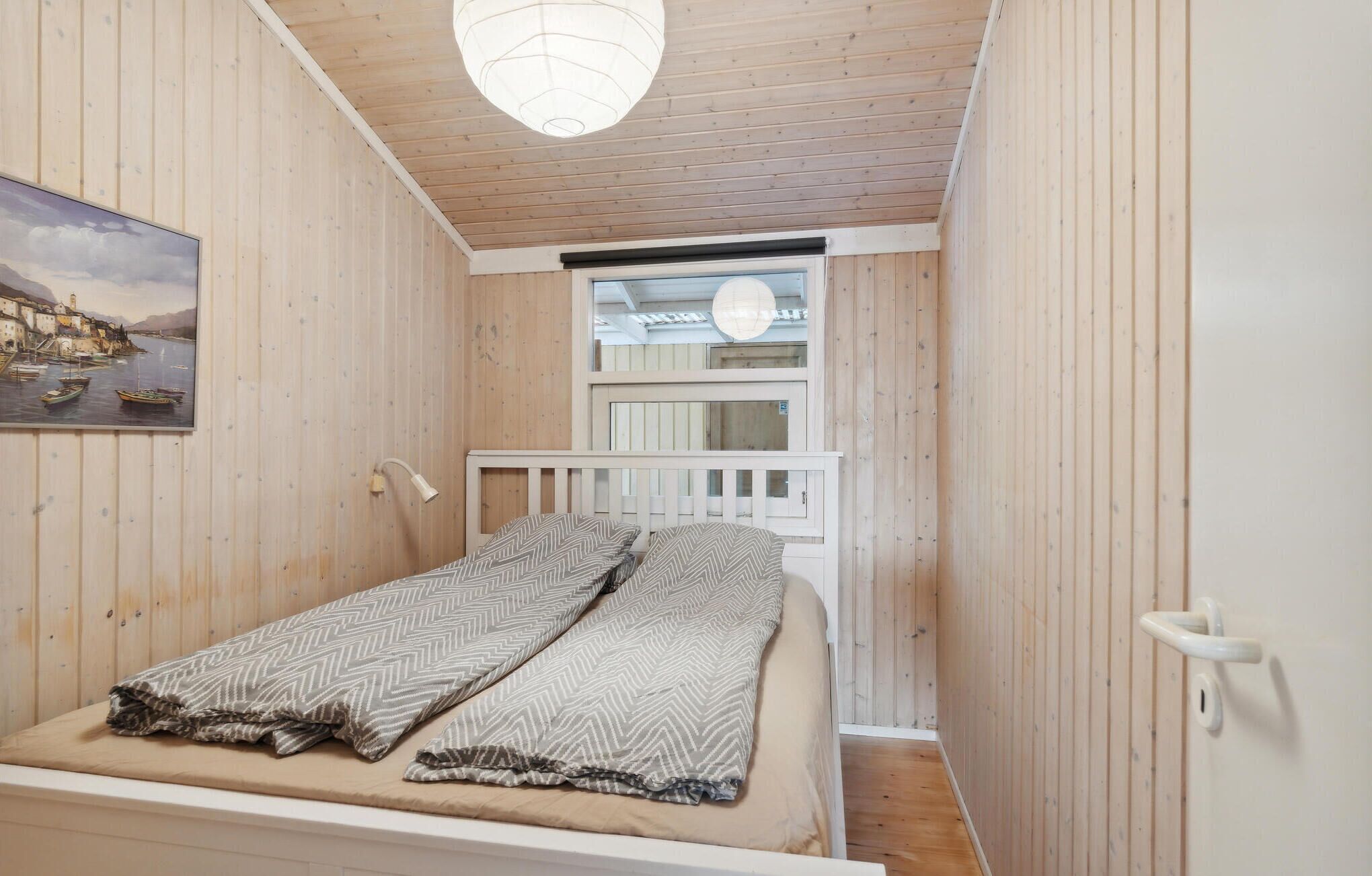 Une petite pièce chaleureuse avec un plafond et des murs en bois, un lit avec une tête de lit blanche, une fenêtre donnant sur la mer et un tableau représentant un paysage côtier.