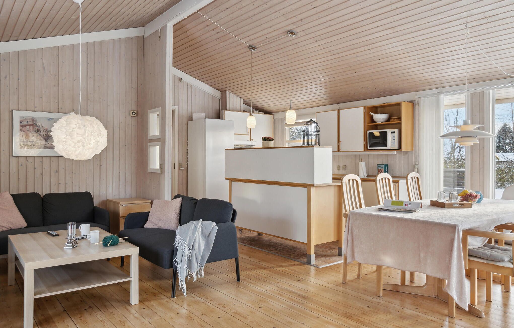 En moderne, åben stue og spiseområde med trægulv, en sofa, et sofabord, et spisebord med stole, og et køkken med skabe og en mikrobølgeovn.