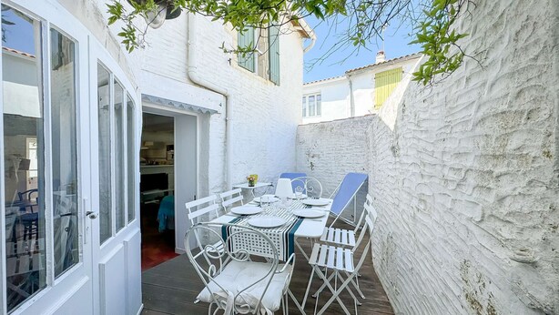 A small outdoor patio with a table set for two, surrounded by white chairs, against a white wall with a window.