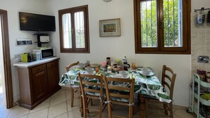 Een eetgedeelte met een tafel voor vier personen, houten stoelen, een keuken met een magnetron en broodrooster, en een raam met uitzicht op groen.