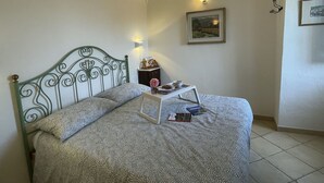 Een bed met een bedsprei met patroon, een nachtkastje met een dienblad, een ingelijst schilderij aan de muur, en een kleine lamp.