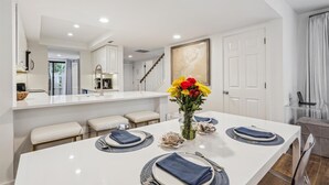 A modern dining area with a white table set for four, a vase of flowers, and a staircase leading to another level. - 112 The Greens @ Shipyard | Golf, Tennis &  Pool | Walk to Beach (Hilton Head Island)
