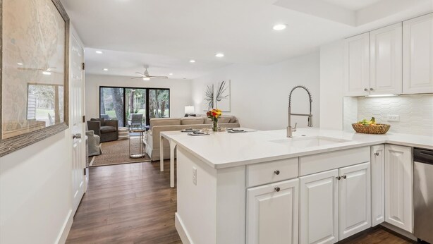 A modern kitchen with white cabinetry, a large island with a sink, and a dining area with a sofa and chairs. - 112 The Greens @ Shipyard | Golf, Tennis &  Pool | Walk to Beach (Hilton Head Island)