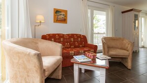 En stue med to beige lænestole, en rød sofa med mønster, et sofabord af glas med magasiner, og et billede på væggen.