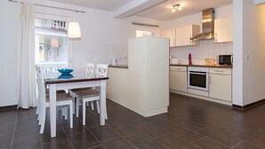 Et moderne køkken med hvide skabe, en indbygget ovn og en mikrobølgeovn. Der er et spiseområde med et hvidt bord og stole, samt en blå skål på bordet.