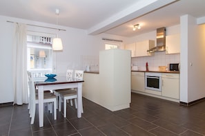 A modern kitchen with white cabinets, a built-in oven, and a microwave. There is a dining area with a white table and chairs, and a blue bowl on the table.
