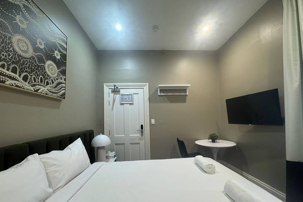 Habitación de hotel con una cama, un escritorio, una silla, una mesita con una planta, un televisor y un cuadro en la pared.