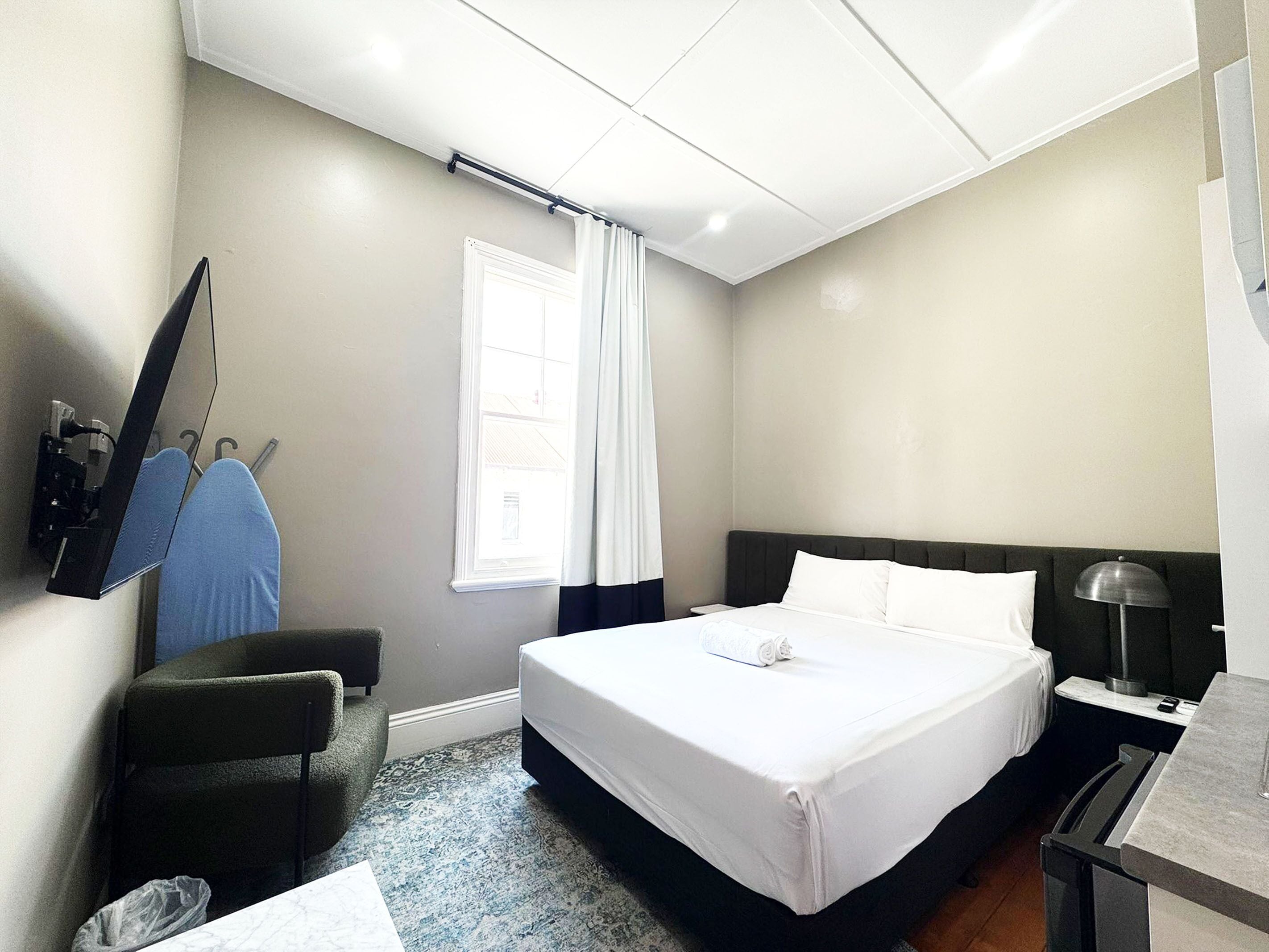 A hotel room with a bed, a desk with a lamp, a chair, a TV mounted on the wall, and a window with curtains.