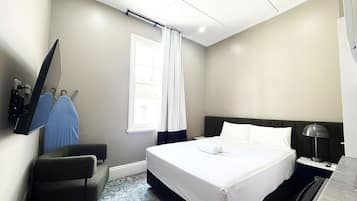 A hotel room with a bed, a desk with a lamp, a chair, a TV mounted on the wall, and a window with curtains.