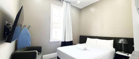 A hotel room with a bed, a desk with a lamp, a chair, a TV mounted on the wall, and a window with curtains.