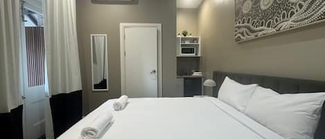 A hotel room with a large bed, a bedside table, a wall-mounted air conditioner, a small kitchenette, and a decorative wall piece.