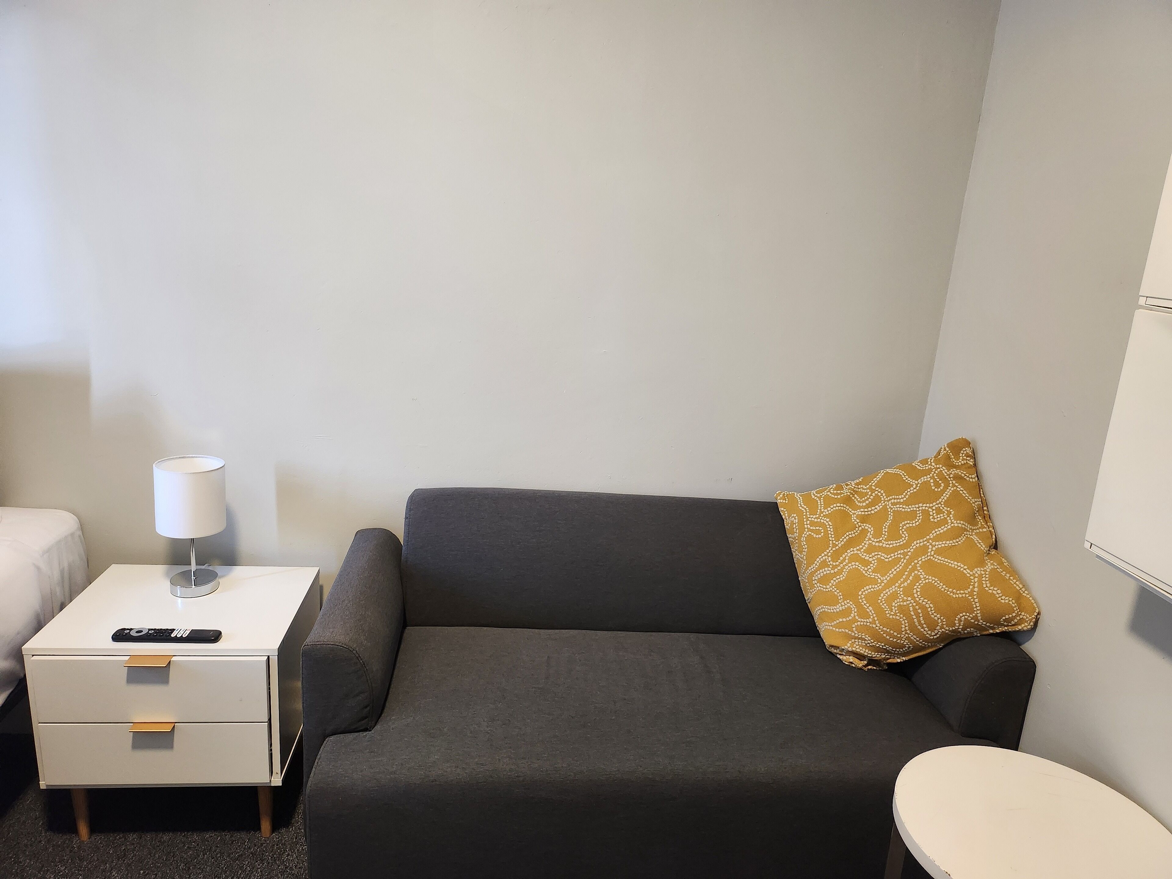 A small, modern hotel room with a grey sofa, a white bedside table with a lamp, a remote control, and a patterned pillow.