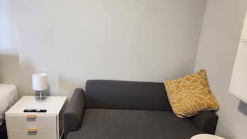 A small, modern hotel room with a grey sofa, a white bedside table with a lamp, a remote control, and a patterned pillow.