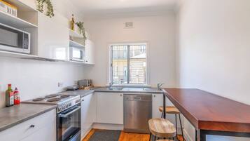 Una cocina compacta equipada con microondas, estufa y lavavajillas. Hay una ventana que proporciona luz natural, y un banco de madera en la encimera.