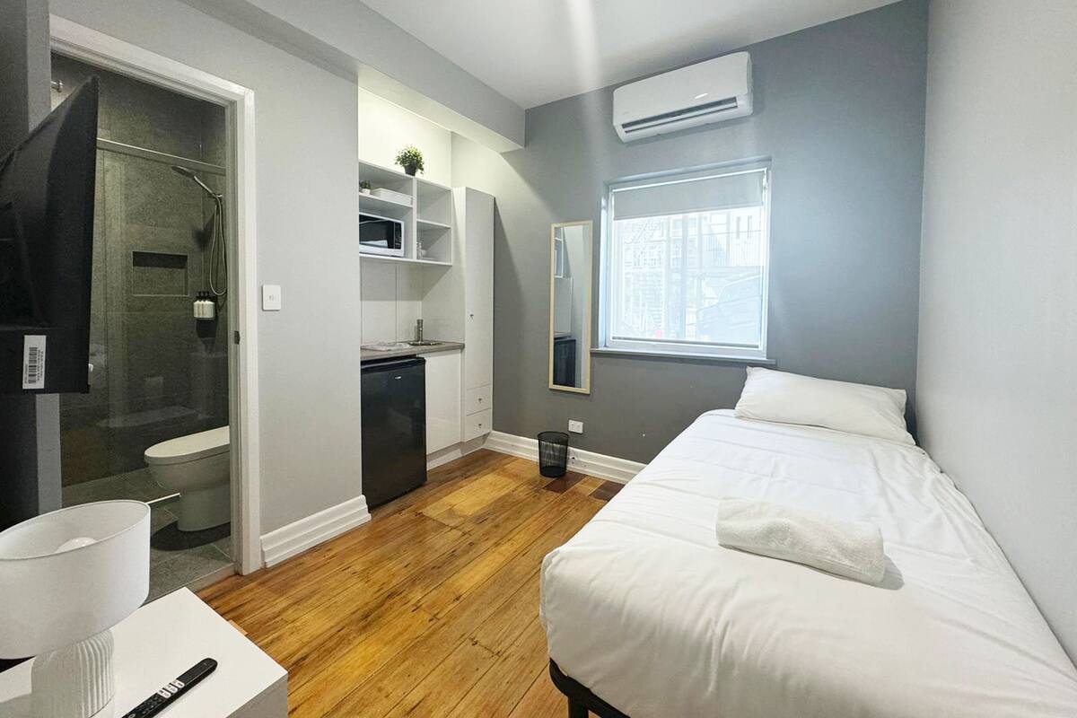 Habitación compacta con cama, mesita de noche, kitchenette pequeño, baño y vestidor.