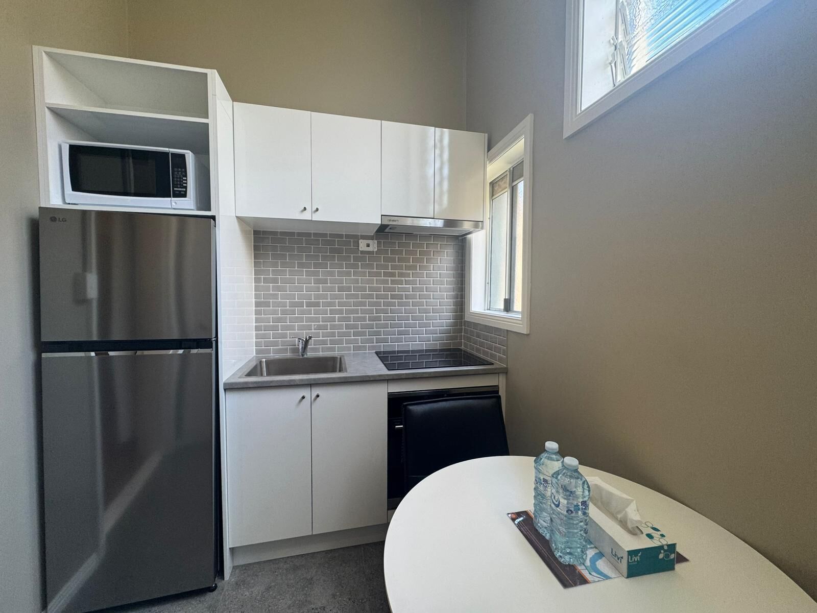 A compact kitchen with white cabinets, a stainless steel refrigerator, a microwave, and a sink. There is a window providing natural light and a table with bottled water and a tissue box.